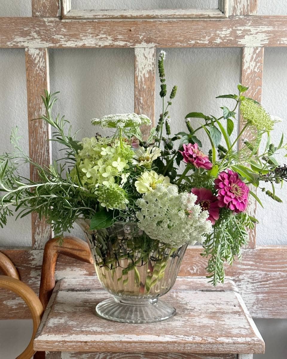A clear glass vase filled with a mixed bouquet of green, white, and bright pink flowers, along with leafy greenery, sits on a rustic wooden stool—perfect for adding charm to any Outdoor Spaces Summer Home Tour.