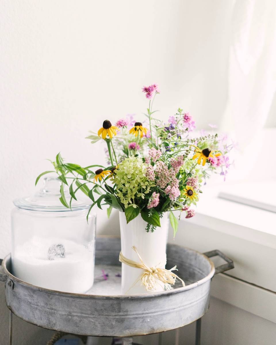A white vase filled with assorted wildflowers sits on a metal tray next to a glass jar of sugar with a scoop, positioned by a window in a bright, airy room—perfect inspiration for your Outdoor Spaces Summer Home Tour.