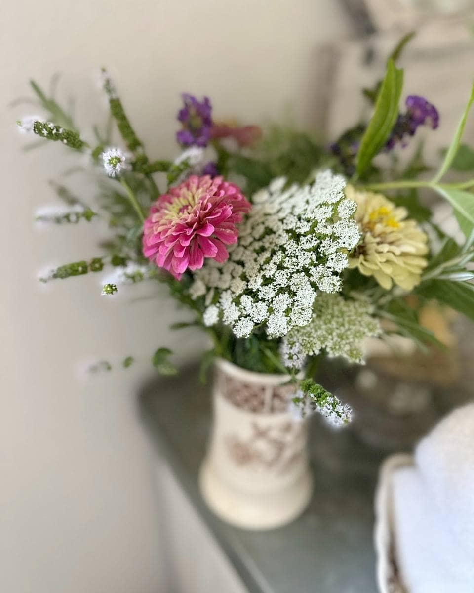 A ceramic vase holds a bouquet of assorted flowers—including a pink zinnia, white lace flowers, and greenery—perfect for adding fresh color to outdoor spaces on your Summer Home Tour. The arrangement sits on a table with a soft, blurred background.