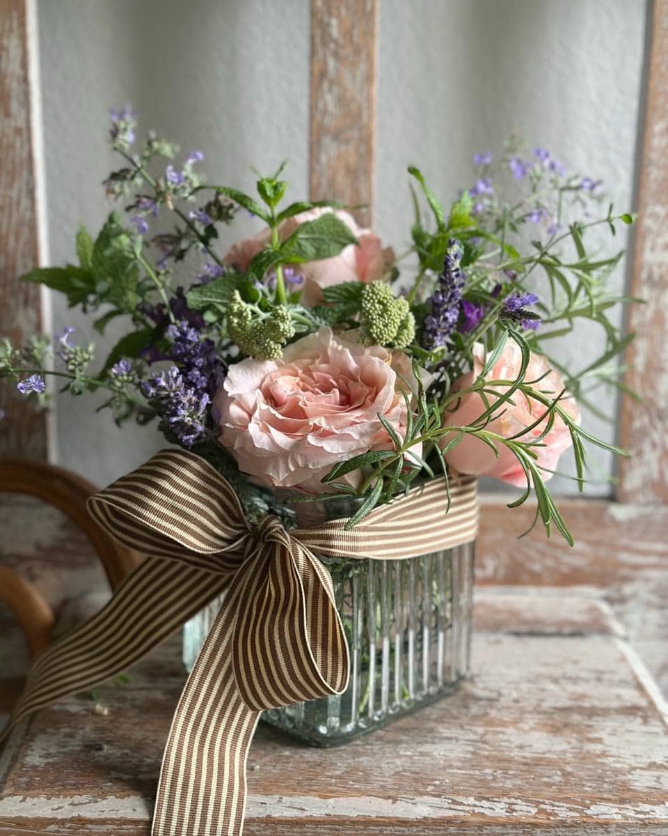 A glass vase with pink roses, purple wildflowers, and greenery, tied with a brown and white striped ribbon, sits on a rustic wooden surface in front of a window—perfect for your Outdoor Spaces Summer Home Tour.