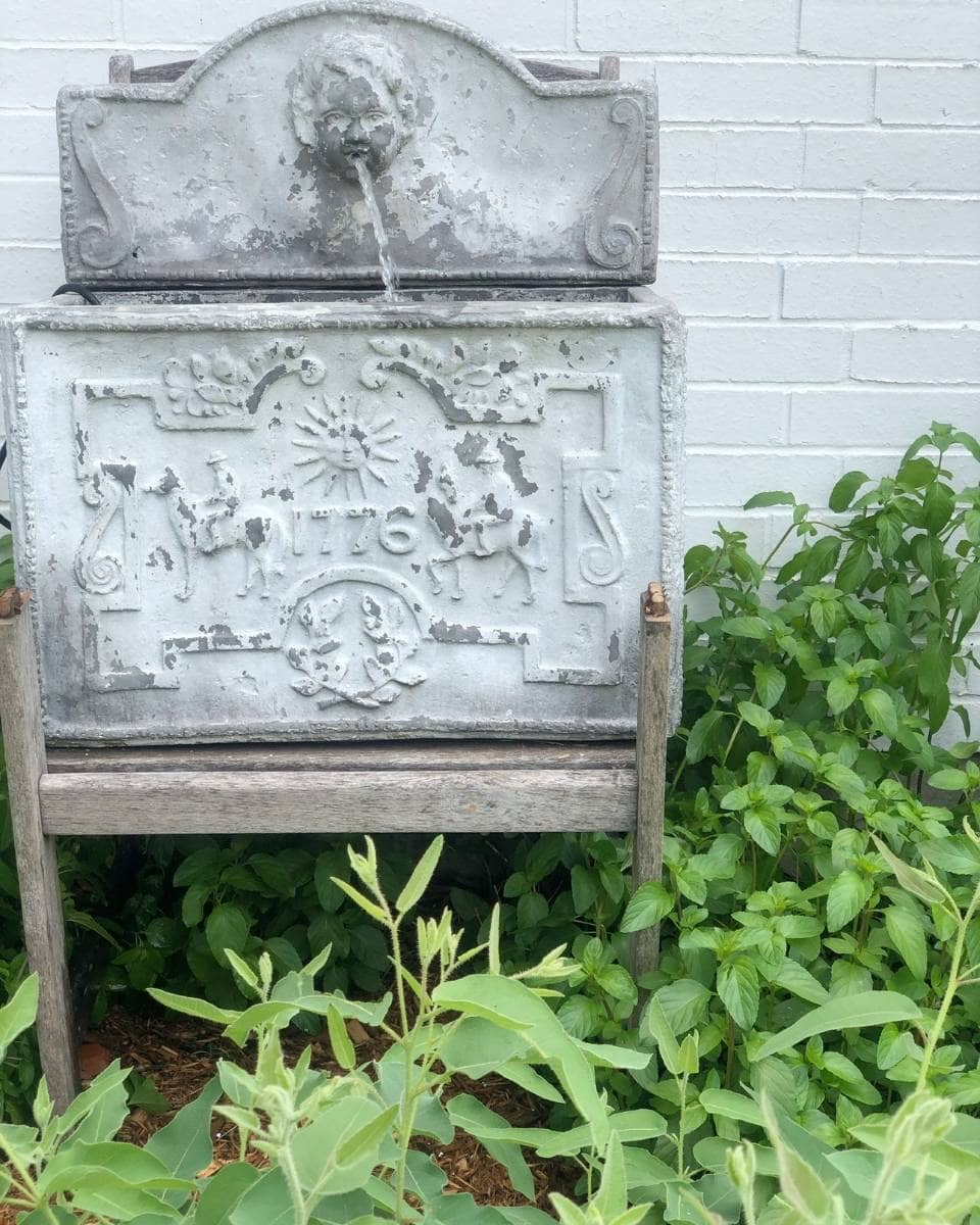 A decorative, weathered stone water fountain with carved designs stands against a white brick wall, surrounded by lush green plants—an inviting feature for Outdoor Spaces on your Summer Home Tour. Water gently flows from the top spout into the basin below.