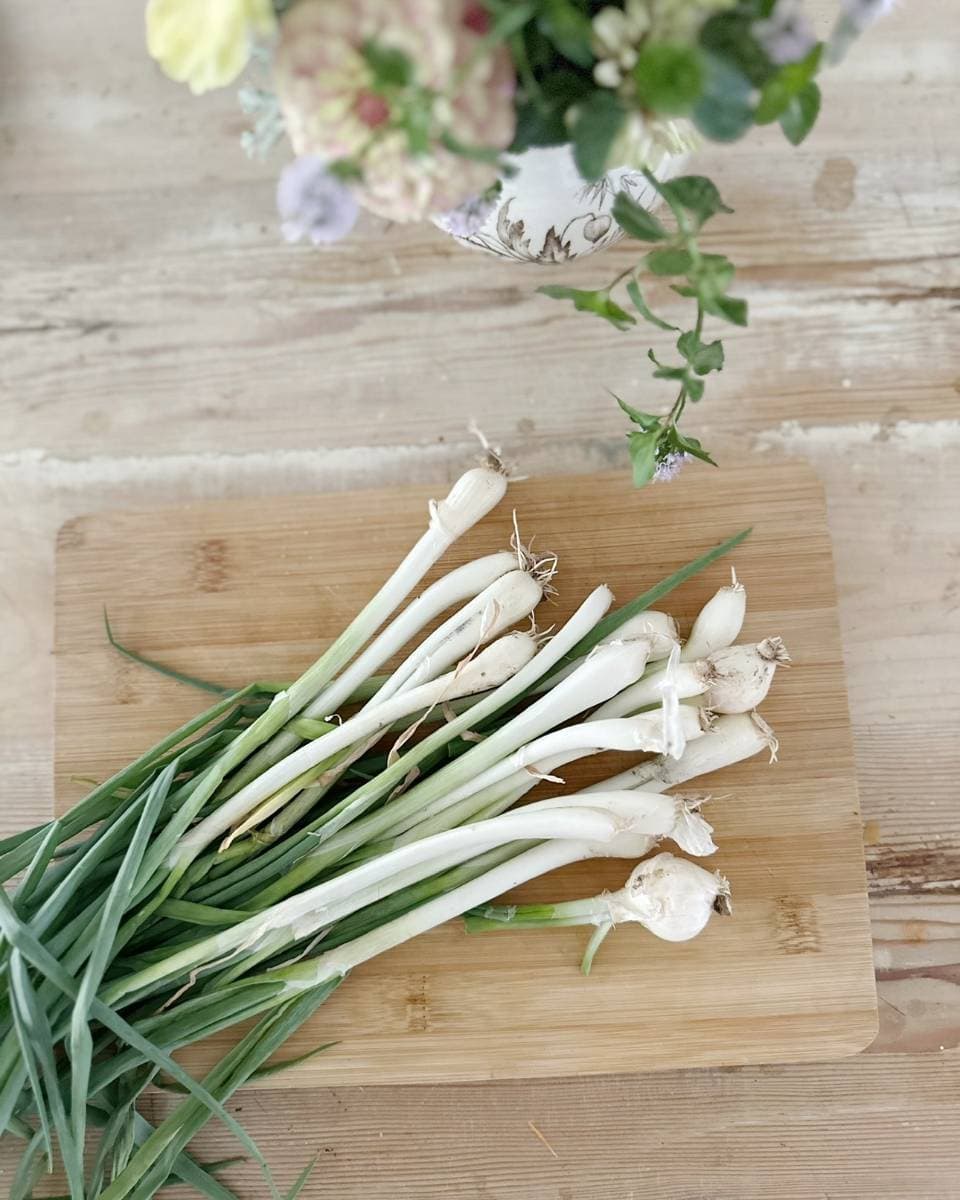 A bunch of fresh green onions with roots attached lies on a wooden cutting board, evoking the vibrant feel of an Outdoor Spaces Summer Home Tour. A floral arrangement in a decorative vase peeks in at the top of the image on a light wooden table.