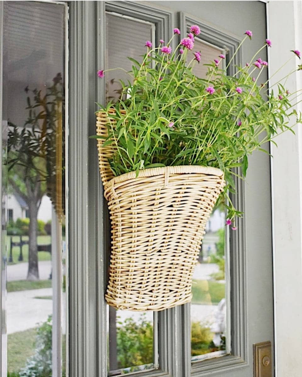 A wicker basket filled with green stems and small purple flowers hangs on a gray front door with glass panels, setting a welcoming scene for any Outdoor Spaces Summer Home Tour. Through the glass, a tree and a house are visible in the background.