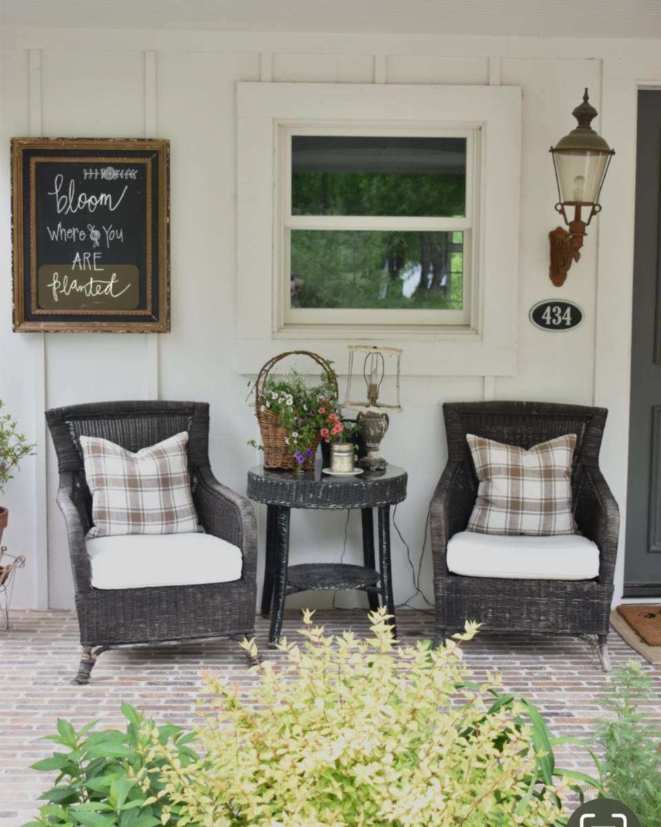A cozy porch with two wicker chairs topped with plaid pillows, a round side table displaying potted plants and decorative items, a wall sign reading "bloom where you are planted," and a lantern sconce by the door—perfect for an Outdoor Spaces Summer Home Tour.