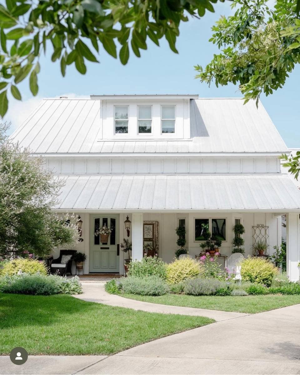 A charming white farmhouse with a metal roof, wide front porch, green door, and lush landscaping invites you to an Outdoor Spaces Summer Home Tour beneath a bright blue sky.