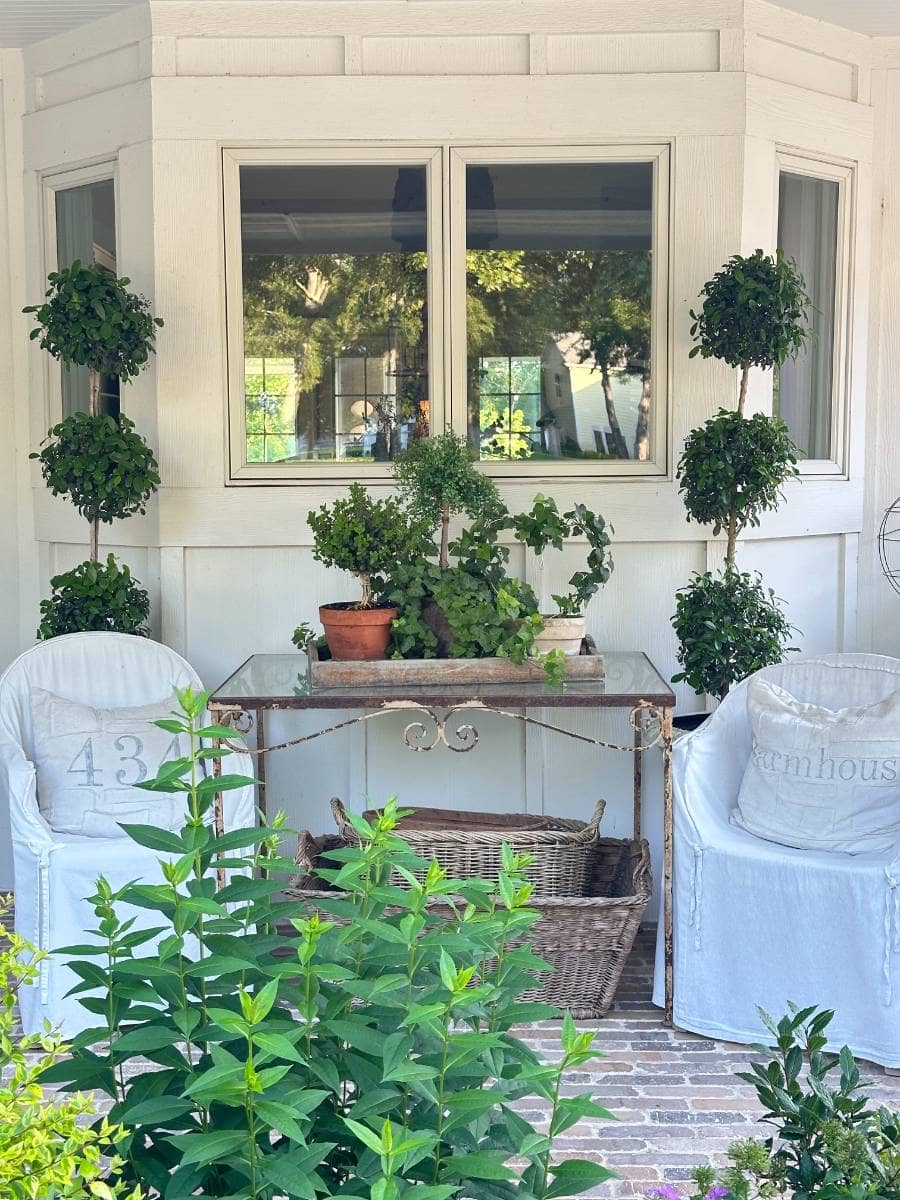 A cozy porch with two white slipcovered chairs, a glass-top table with potted plants, and topiary trees captures the charm of Outdoor Spaces. This light farmhouse Summer Home Tour features greenery, wicker accents, and large windows in the background.