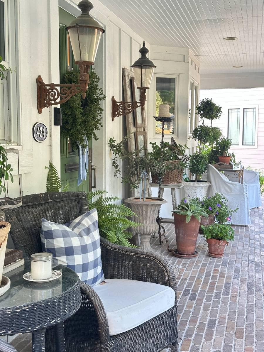 A cozy porch with wicker chairs, a blue plaid pillow, potted plants, and lantern-style lights sets the stage for an inviting Outdoor Spaces Summer Home Tour. A white candle sits on the glass-topped table beside the chair, completing this peaceful scene.
