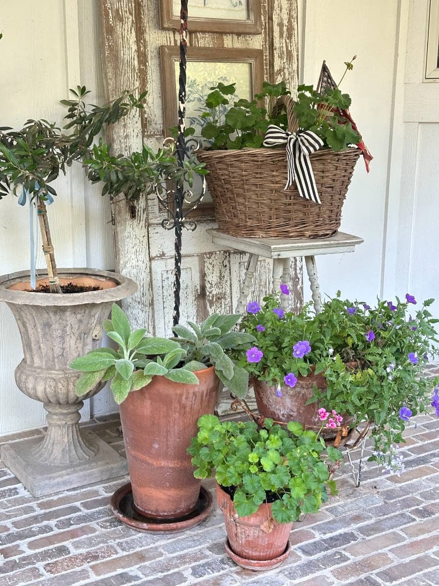 Several potted plants and flowers in clay pots and a wicker basket are arranged on a brick porch in front of a rustic wooden door, creating a charming, vintage display perfect for an Outdoor Spaces Summer Home Tour.