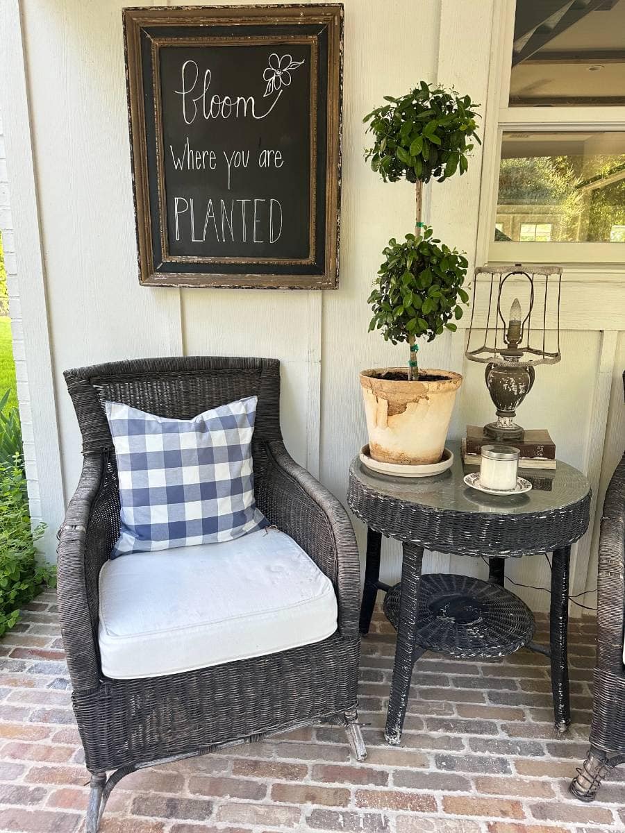 A cozy porch scene from an Outdoor Spaces Summer Home Tour: a wicker chair topped with a blue and white checkered pillow, a round side table with a potted plant, candle, lamp, and a chalkboard sign reading, "Bloom where you are planted.
