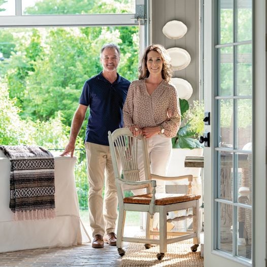 A smiling man and woman stand together in a bright, sunlit room with large windows, inviting Outdoor Spaces inside. Light-colored furniture, plants, and decorative plates on the wall create an airy Summer Home Tour atmosphere.