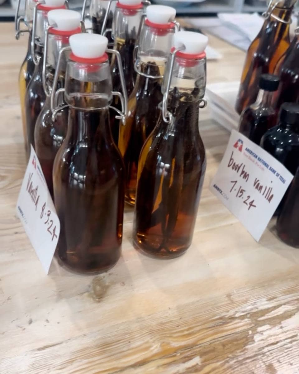 Several glass bottles with swing-top lids, filled with dark liquid, are arranged on a wooden table. White labels read "Vanilla 8-3-24" and "Bourbon Vanilla 7-15-24," showcasing an easy homemade vanilla extract DIY project.