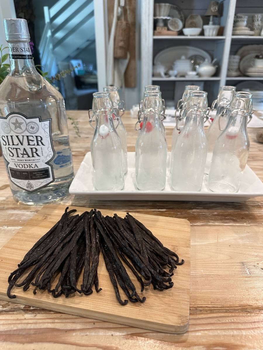 A bottle of Silver Star vodka, six clear glass bottles with stoppers, and several vanilla beans on a wooden board are arranged on a wooden table, perfect for an Easy Homemade Vanilla Extract DIY in a cozy kitchen setting.