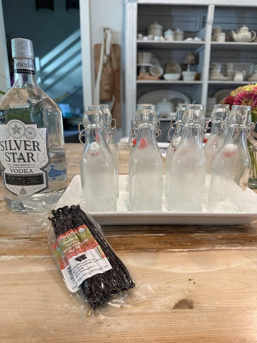 A bottle of Silver Star vodka, six glass bottles with liquid, and a pack of vanilla beans sit on a wooden table—everything you need for Easy Homemade Vanilla Extract DIY. Shelves with dishes and a vase of flowers are in the background.
