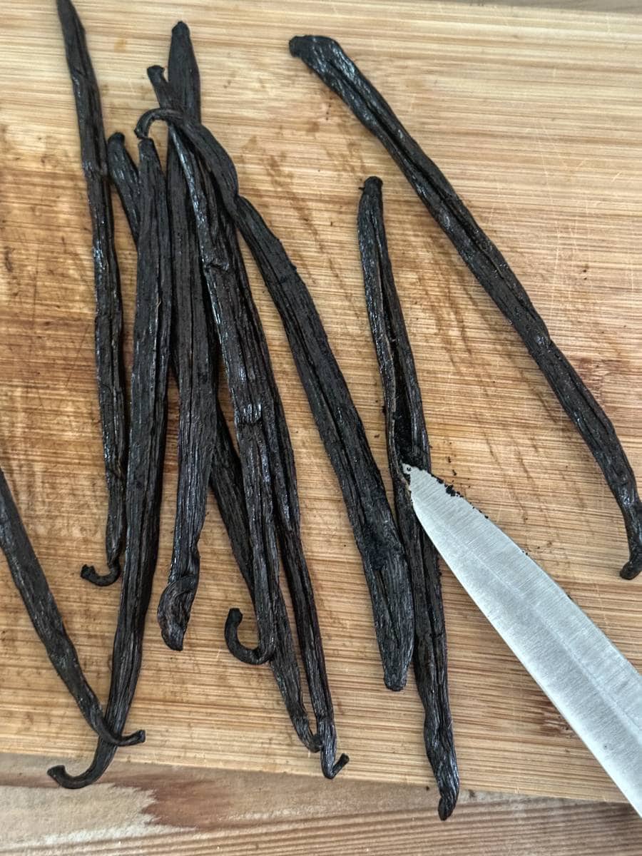 Several dried vanilla beans are laid out on a wooden cutting board, ready for easy homemade vanilla extract DIY. A knife rests nearby, its tip pointing toward one of the beans. The cutting board surface shows marks from regular use.