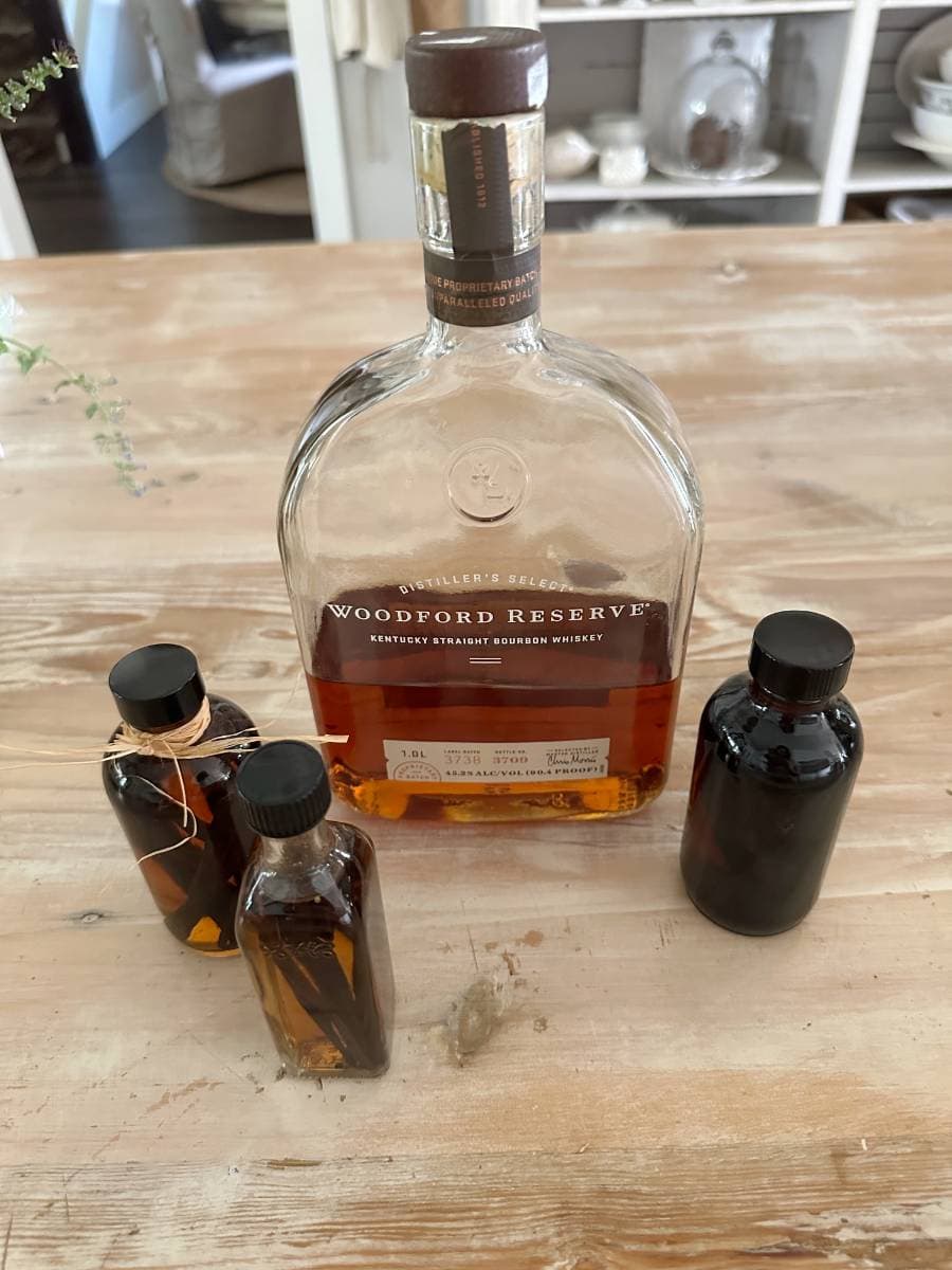 A bottle of Woodford Reserve Kentucky Straight Bourbon Whiskey sits on a wooden table, surrounded by three smaller bottles of Easy Homemade Vanilla Extract DIY. A blurred background reveals home decor on shelves.