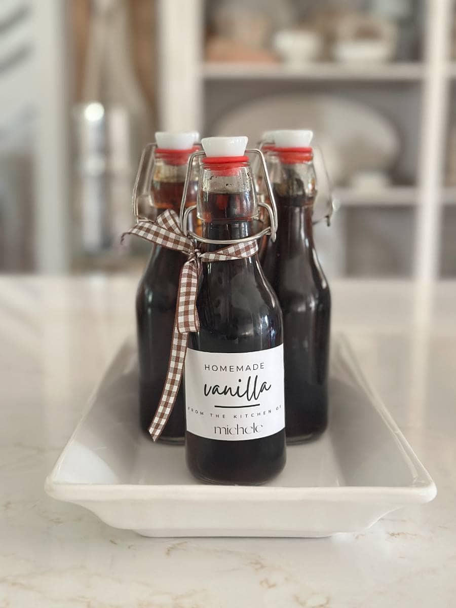 Three glass bottles of easy homemade vanilla extract with clasp lids sit on a white tray. The front bottle, tied with a brown and white checked ribbon, features a label that reads "Homemade vanilla from the kitchen of Michele—perfect for your next DIY project.