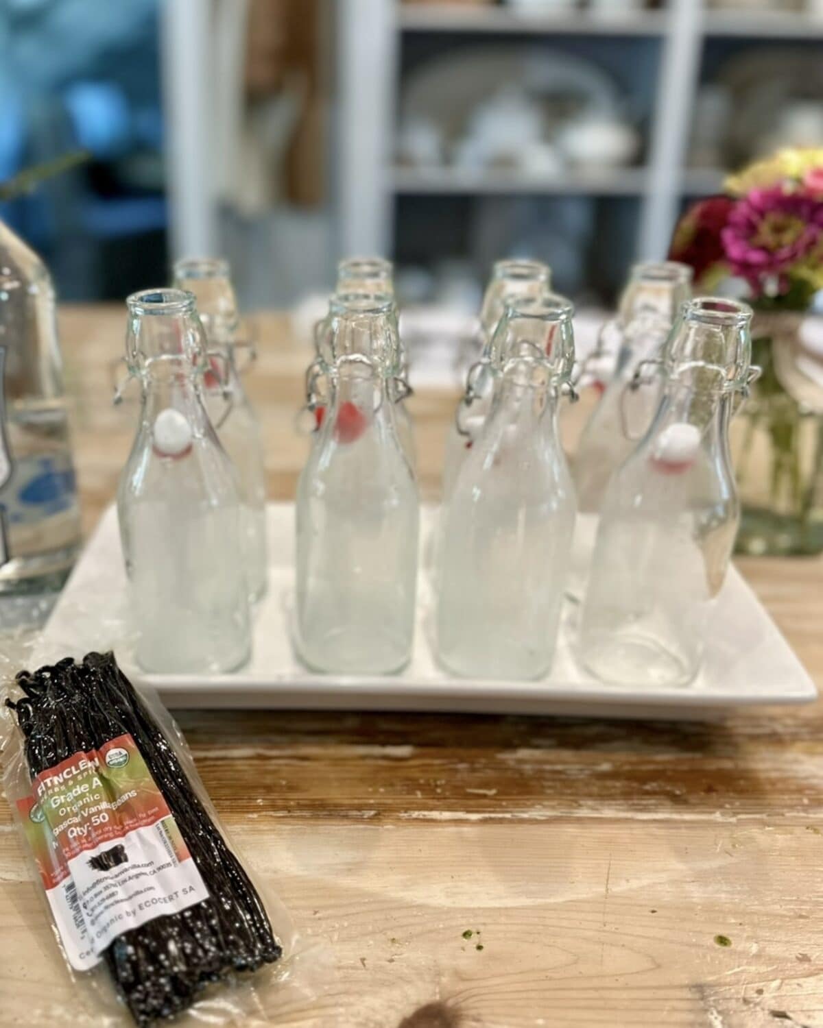 Six empty glass bottles with swing-top lids are arranged on a white tray, ready for Easy Homemade Vanilla Extract DIY. In the foreground, a package of organic vanilla beans rests on a wooden table, with blurred shelves and flowers in the background.