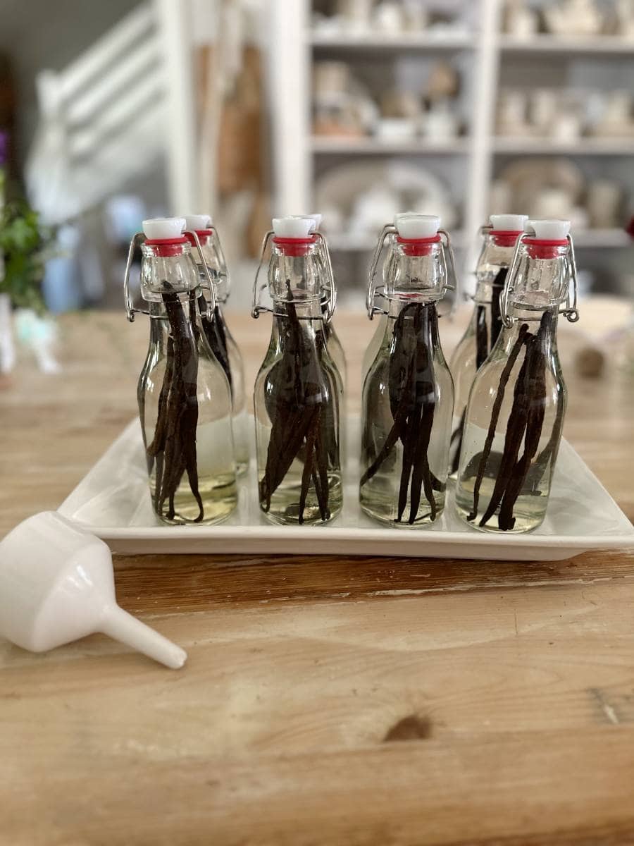 Six glass bottles with swing-top lids, filled with vanilla beans and liquid for Easy Homemade Vanilla Extract DIY, are arranged on a white tray on a wooden table. A white funnel lies in the foreground, with shelves visible in the blurred background.