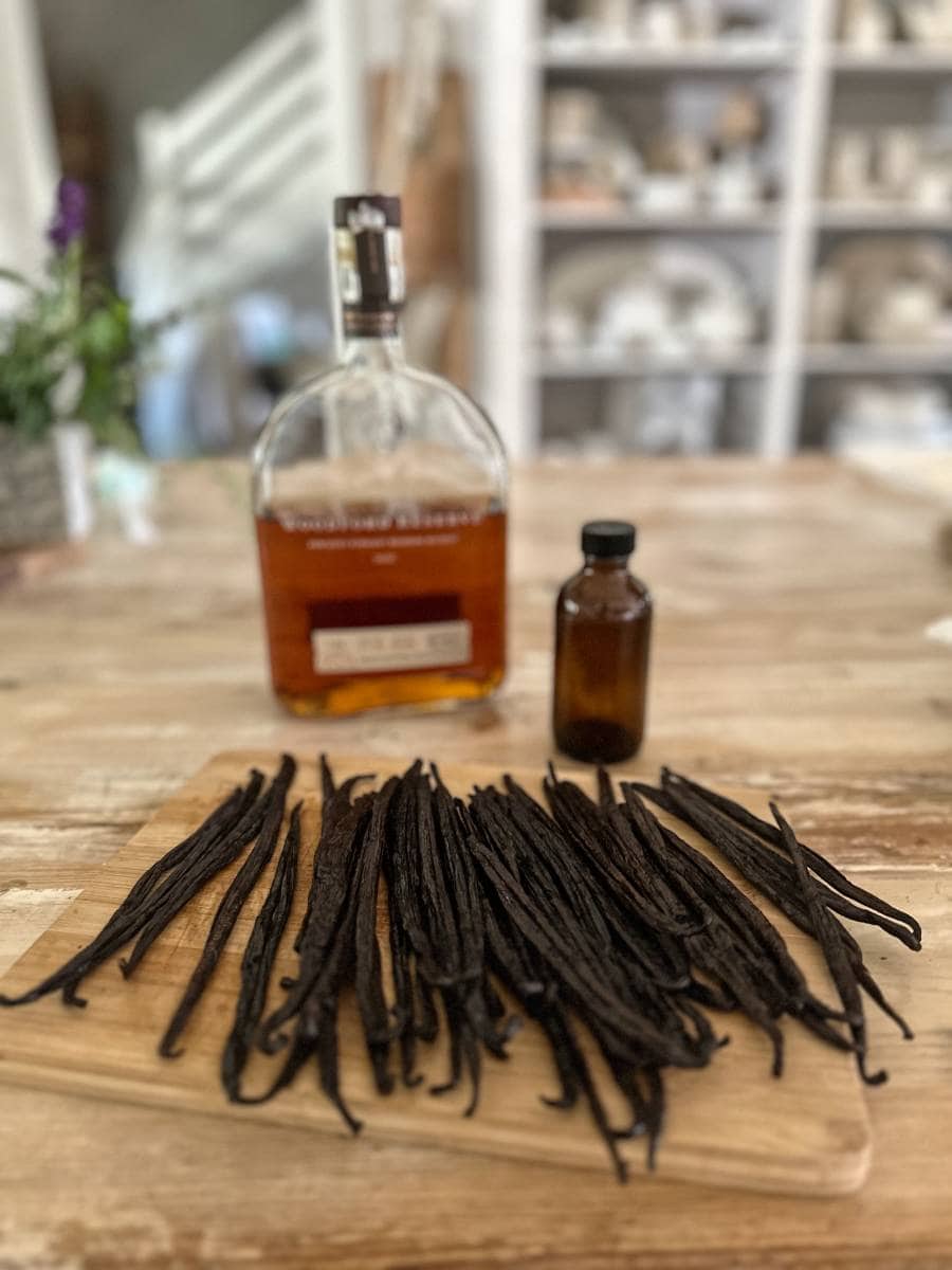 A wooden board with dried vanilla beans sits on a table—perfect for an Easy Homemade Vanilla Extract DIY. Behind it are a bottle of liquor and a small brown bottle, with shelves of dishes blurred in the background.