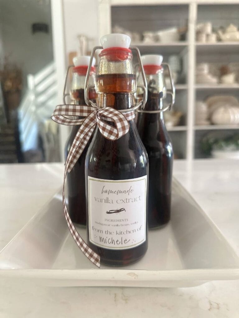 Three glass bottles of easy homemade vanilla extract DIY, adorned with checkered ribbon bows and personalized labels, sit on a white tray on a kitchen counter. Shelves with dishes are visible in the blurred background.