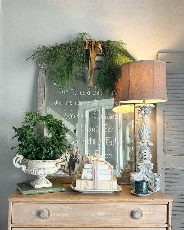 A rustic dresser holds a potted plant, stacked books tied with ribbon, and a decorative lamp. Behind them is a vintage mirror with a Christmas Bible verse written on it and pine garland draped over the top.