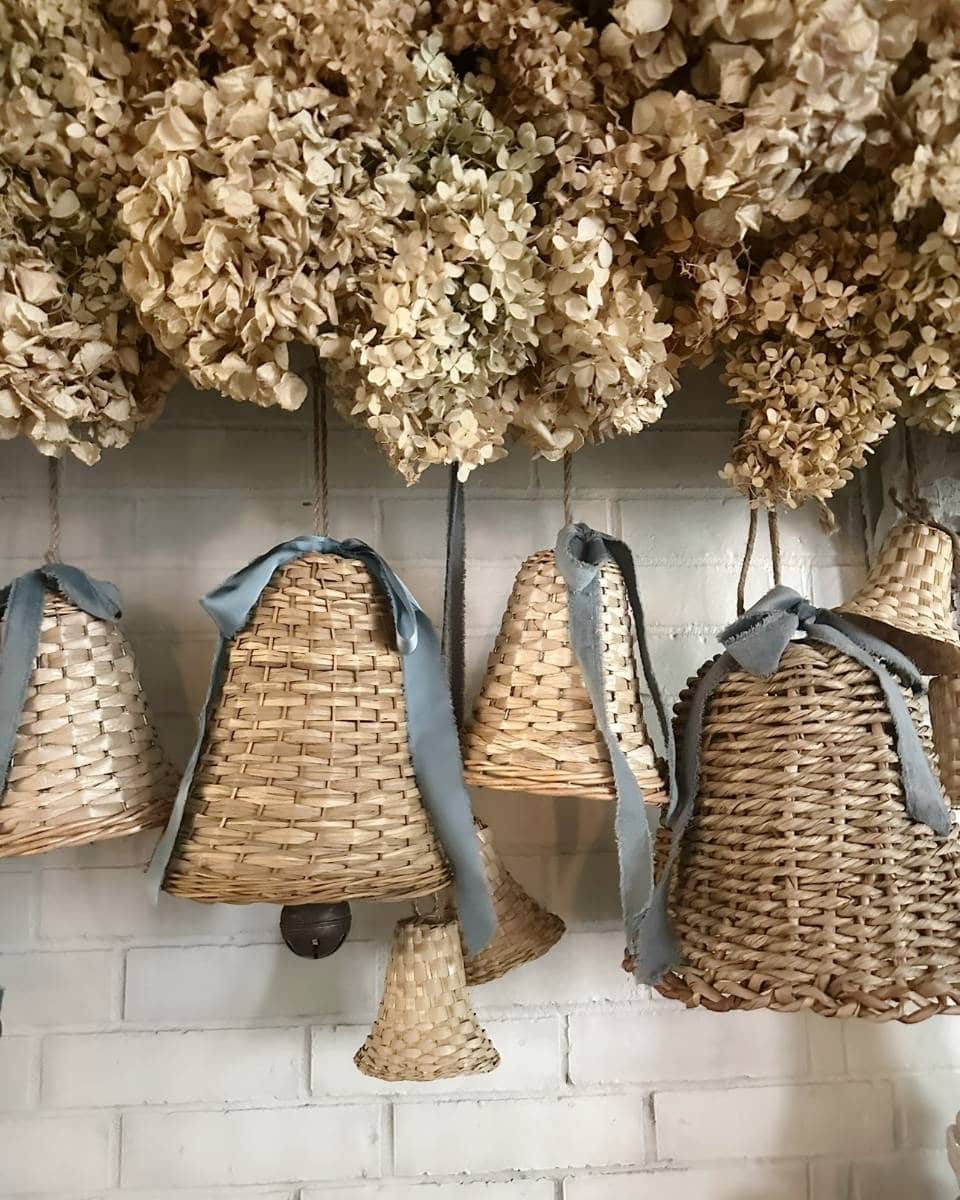 Wicker bells with blue ribbons hang against a white brick wall, with bunches of dried hydrangea flowers arranged above them.