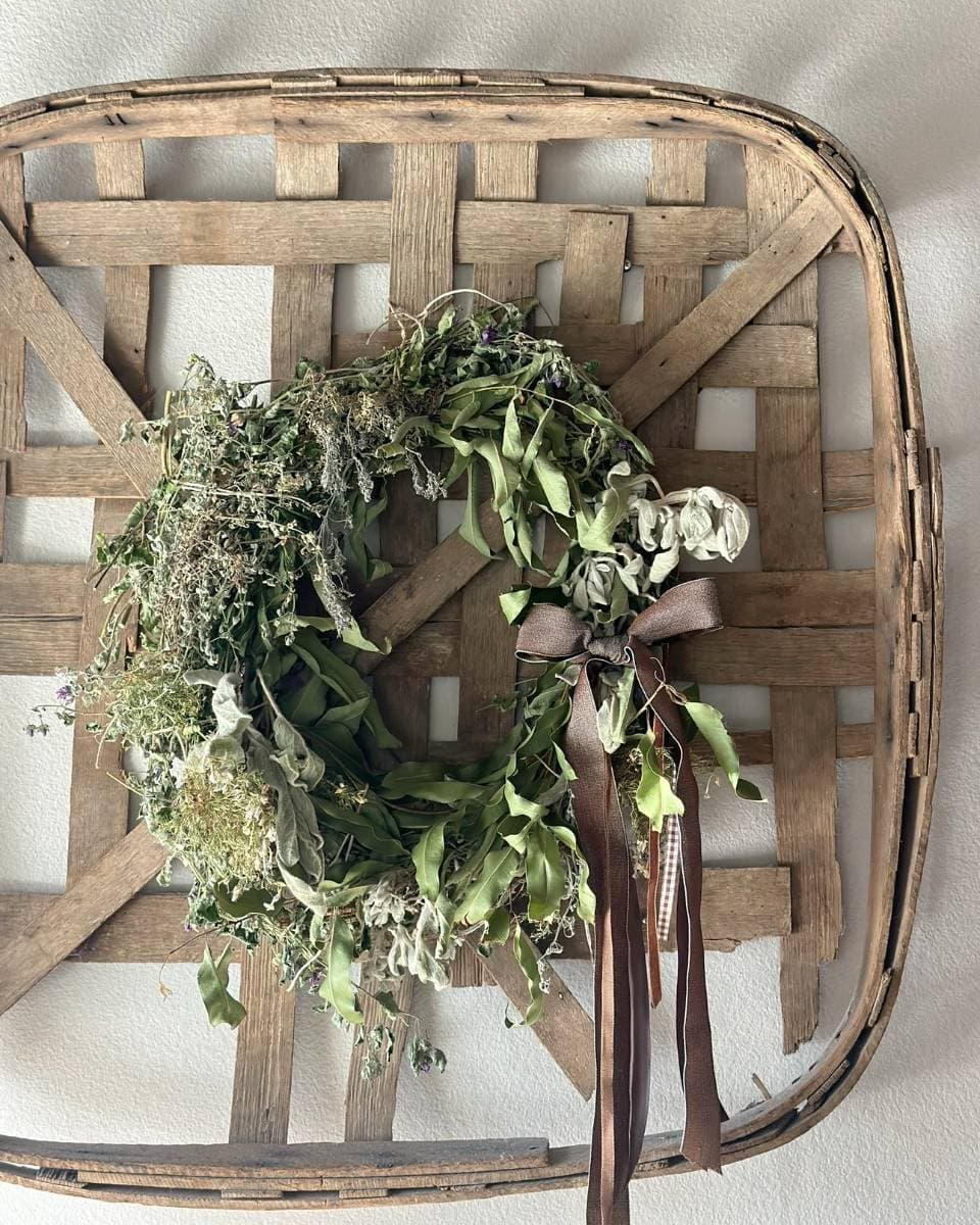 A dried green wreath with a dark brown ribbon is displayed on a rustic woven wooden tray, hanging on a light-colored wall.