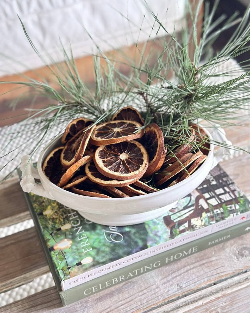 A white bowl filled with dried citrus slices and pine branches sits on top of two books, one titled "CELEBRATING HOME," on a wooden surface with a light, cozy background.