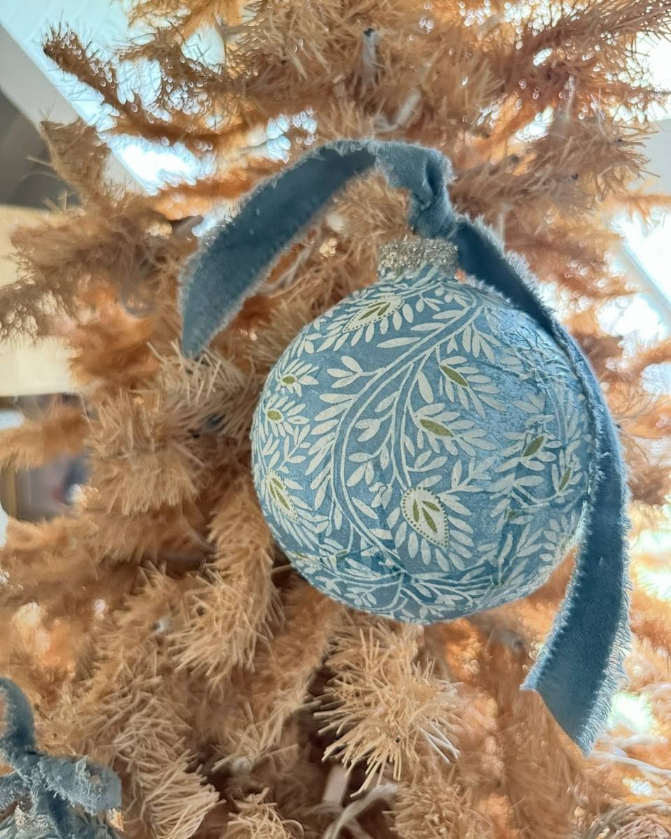 A blue ornament with a leafy white and yellow pattern and a blue velvet ribbon hangs on a dried, beige Christmas tree with soft sunlight in the background.