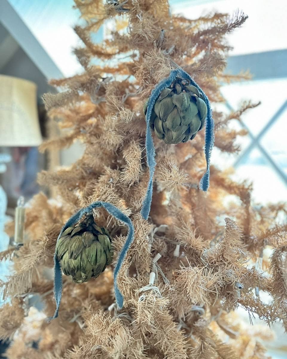 A close-up of a light brown, dried Christmas tree decorated with two hanging artichokes tied with blue velvet ribbons as ornaments. The background is softly blurred, highlighting the unique decorations.