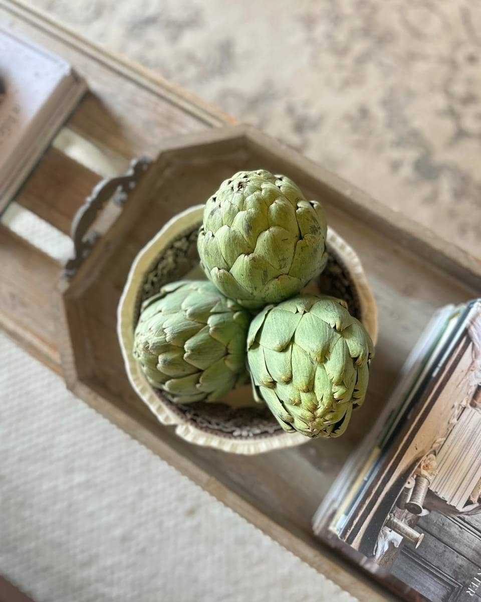 A top-down view of three fresh artichokes in a woven basket on a wooden tray, next to a stack of books and a textured rug underneath.