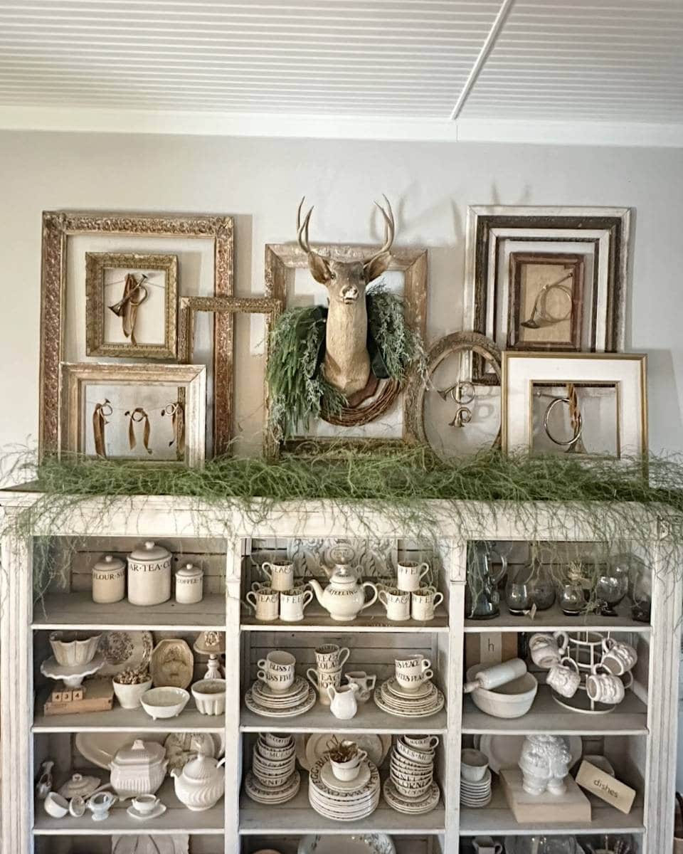 A decorative shelf holds vintage-style cream-colored crockery and teacups. On top, empty ornate frames, a deer head with antlers draped in greenery, and sprigs of foliage create a rustic, eclectic display.