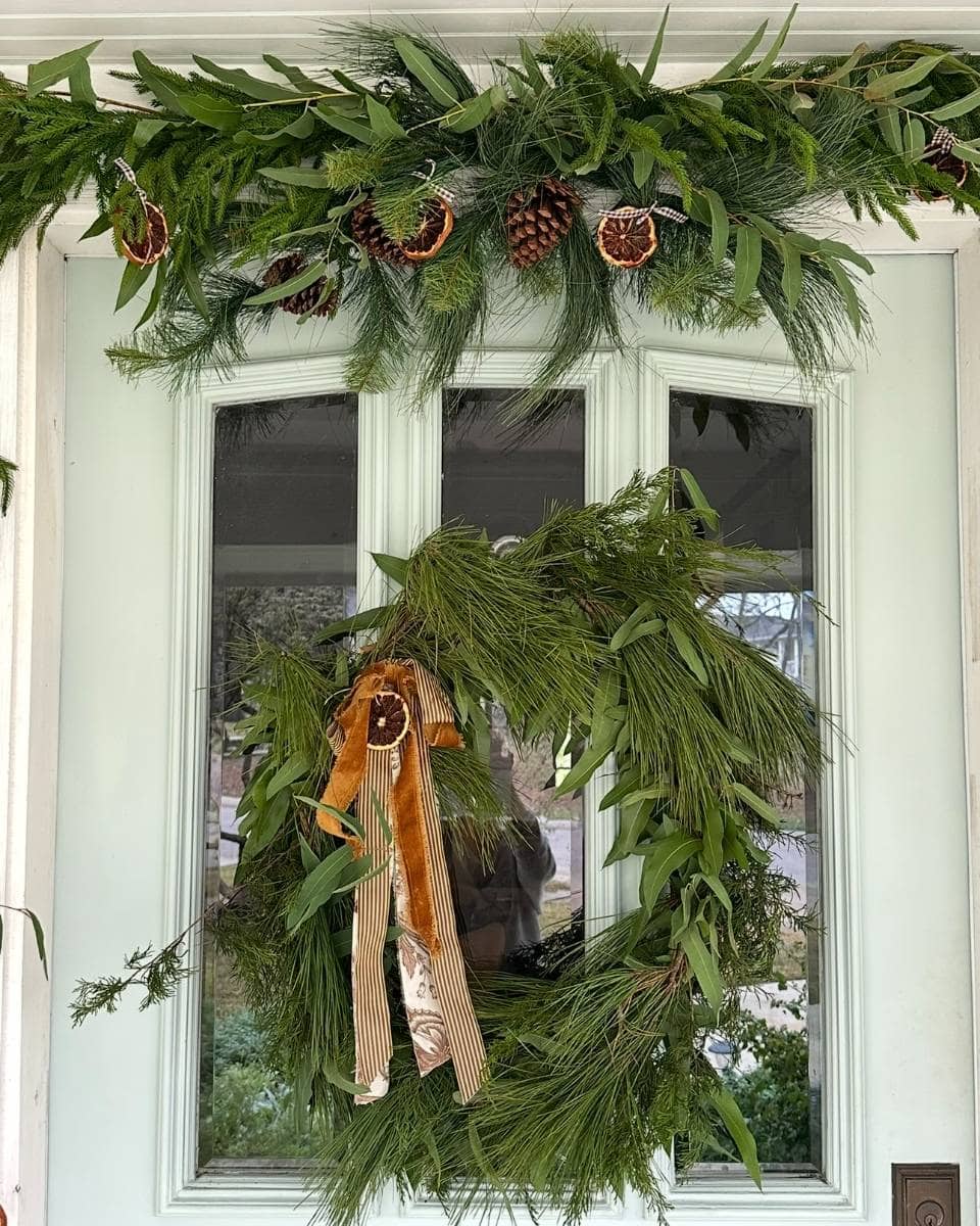 A white door decorated with a green pine wreath featuring a gold ribbon and dried orange slice, and a garland of pine branches, pinecones, and dried oranges above the door.