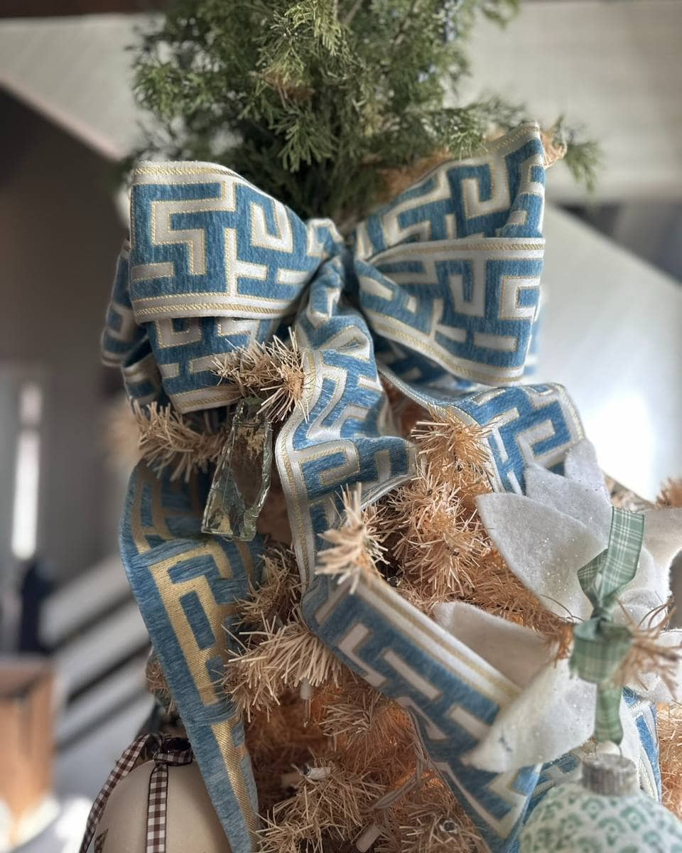 A close-up of a blue and beige patterned ribbon bow tied to the top of a Christmas tree, with white decorative flowers and green foliage in the background.