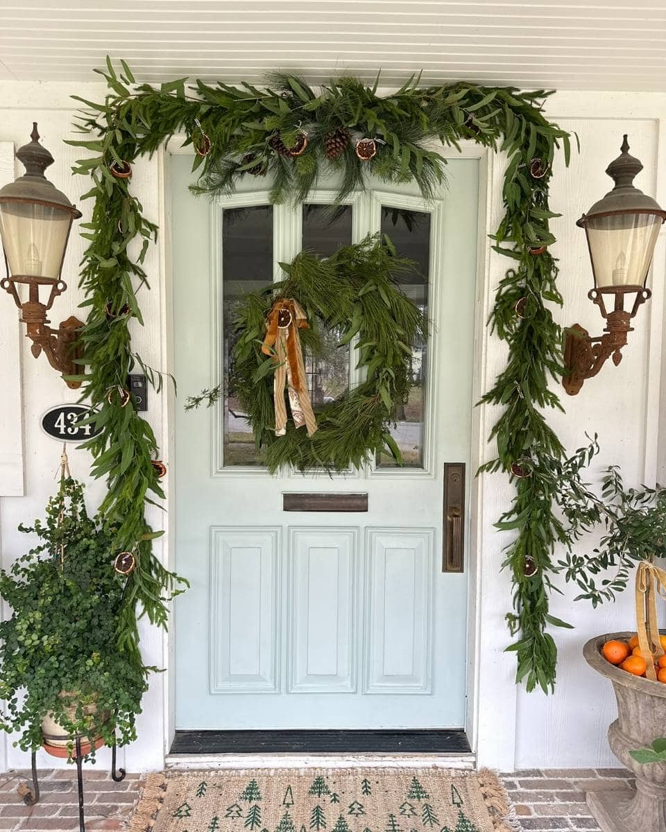 A light blue front door decorated with green pine garland and a wreath, both adorned with dried orange slices. Lanterns hang on each side, and potted plants and a doormat complete the festive entryway.