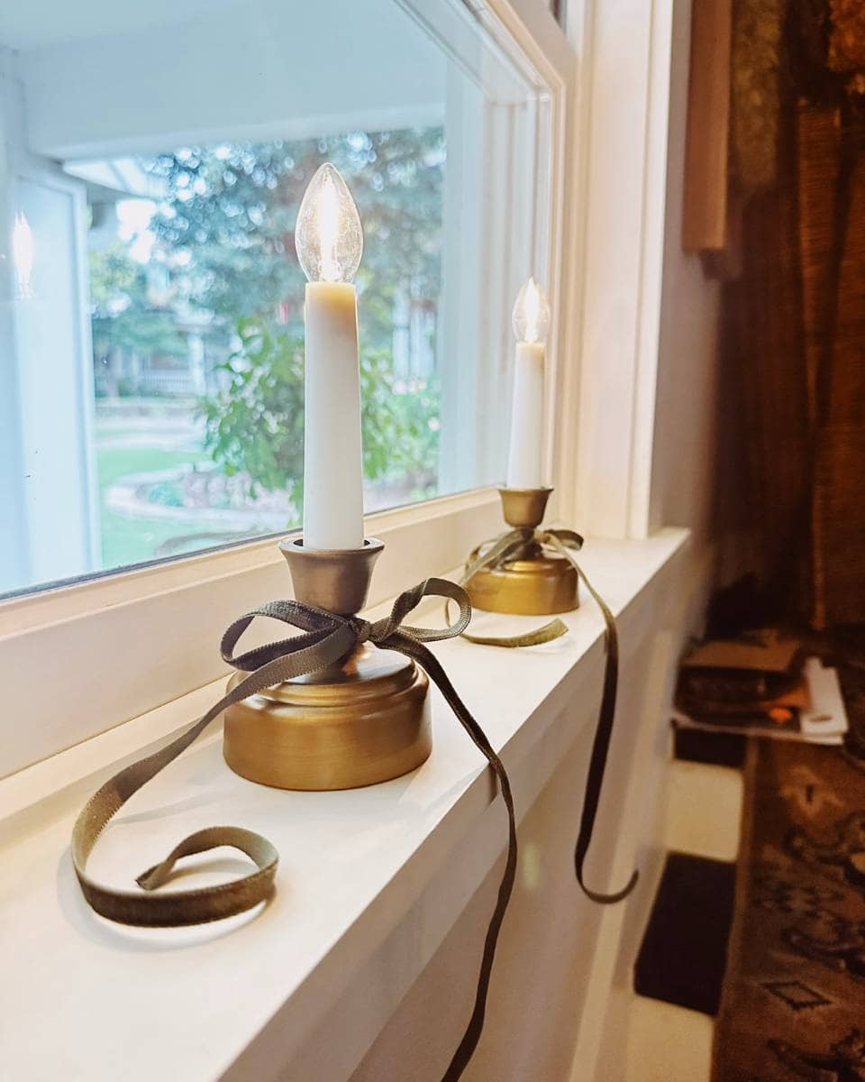 Two electric candles with brass bases and tied with dark ribbon sit on a white windowsill. A garden and trees are visible outside the window, and part of a patterned rug is seen indoors.
