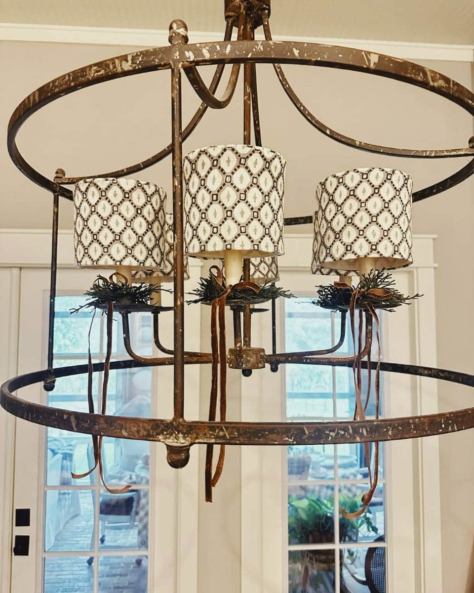 A rustic metal chandelier with three candle-shaped light bulbs, each covered by a patterned white and black lampshade, hangs indoors near glass doors. Greenery and brown ribbon decorates the base of each bulb.
