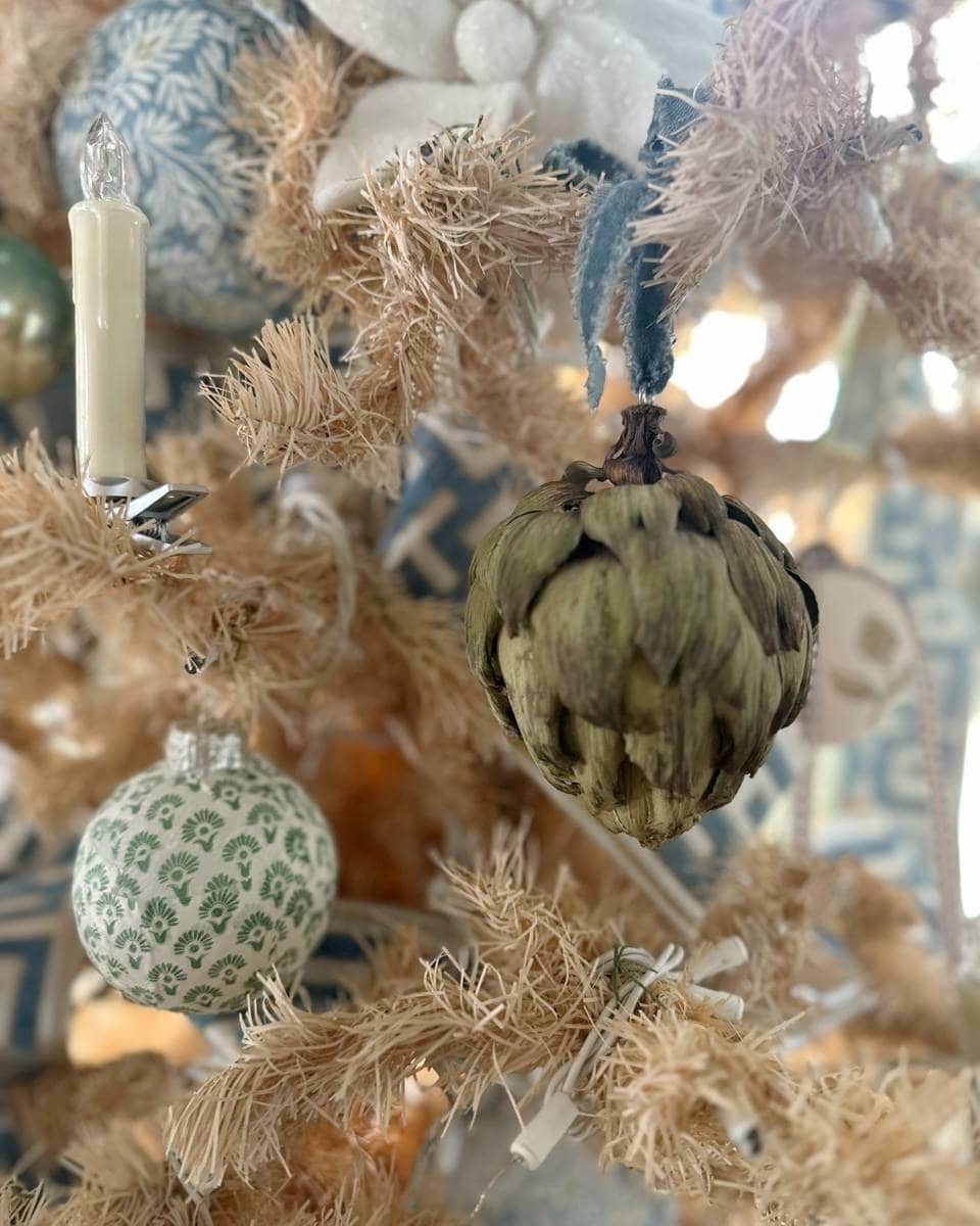 A close-up of a beige Christmas tree decorated with a green artichoke ornament, a patterned round ornament, a white flower, a candle-shaped light, and blue and white ribbon.