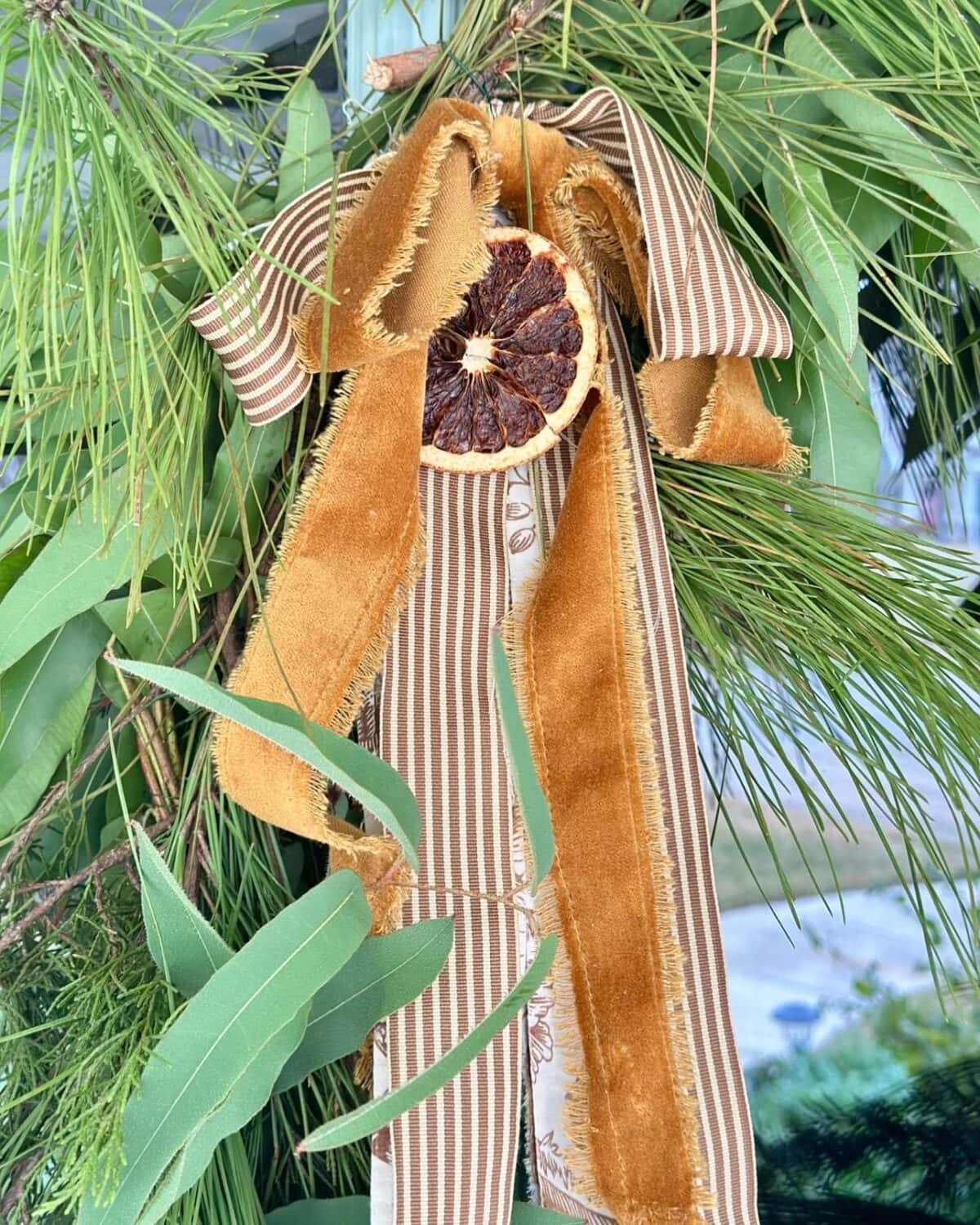 A natural, handmade holiday wreath featuring long pine needles, eucalyptus leaves, a striped brown and cream ribbon, and a dried orange slice—perfect for adding vintage charm to your Christmas tour.