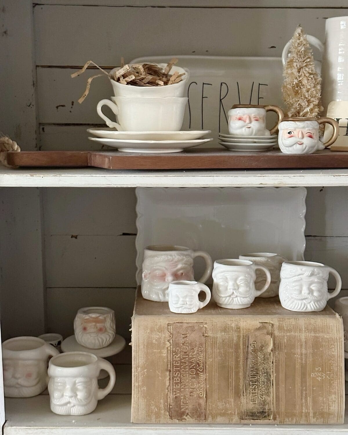 A shelf with natural, handmade ceramic face mugs and cups, including vintage Santa Claus designs, displayed alongside stacked books, plates, and rustic decor—perfect for a cozy Christmas tour in a neutral color palette.