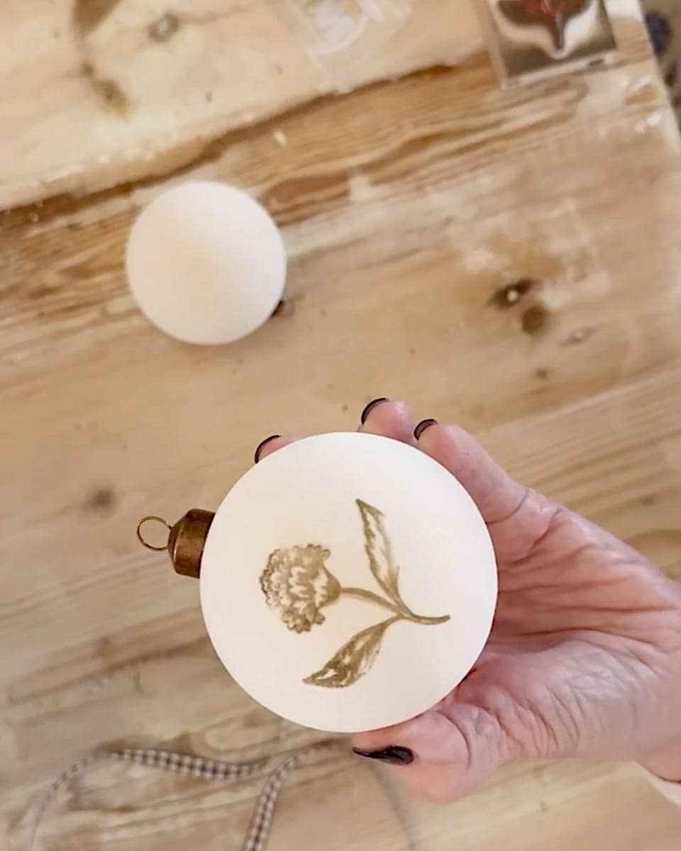 A hand with black nail polish holds a white Christmas ornament decorated with a gold flower illustration. Another plain white ornament and a ribbon rest on a wooden surface, adding a touch of natural vintage charm to your Christmas tour.