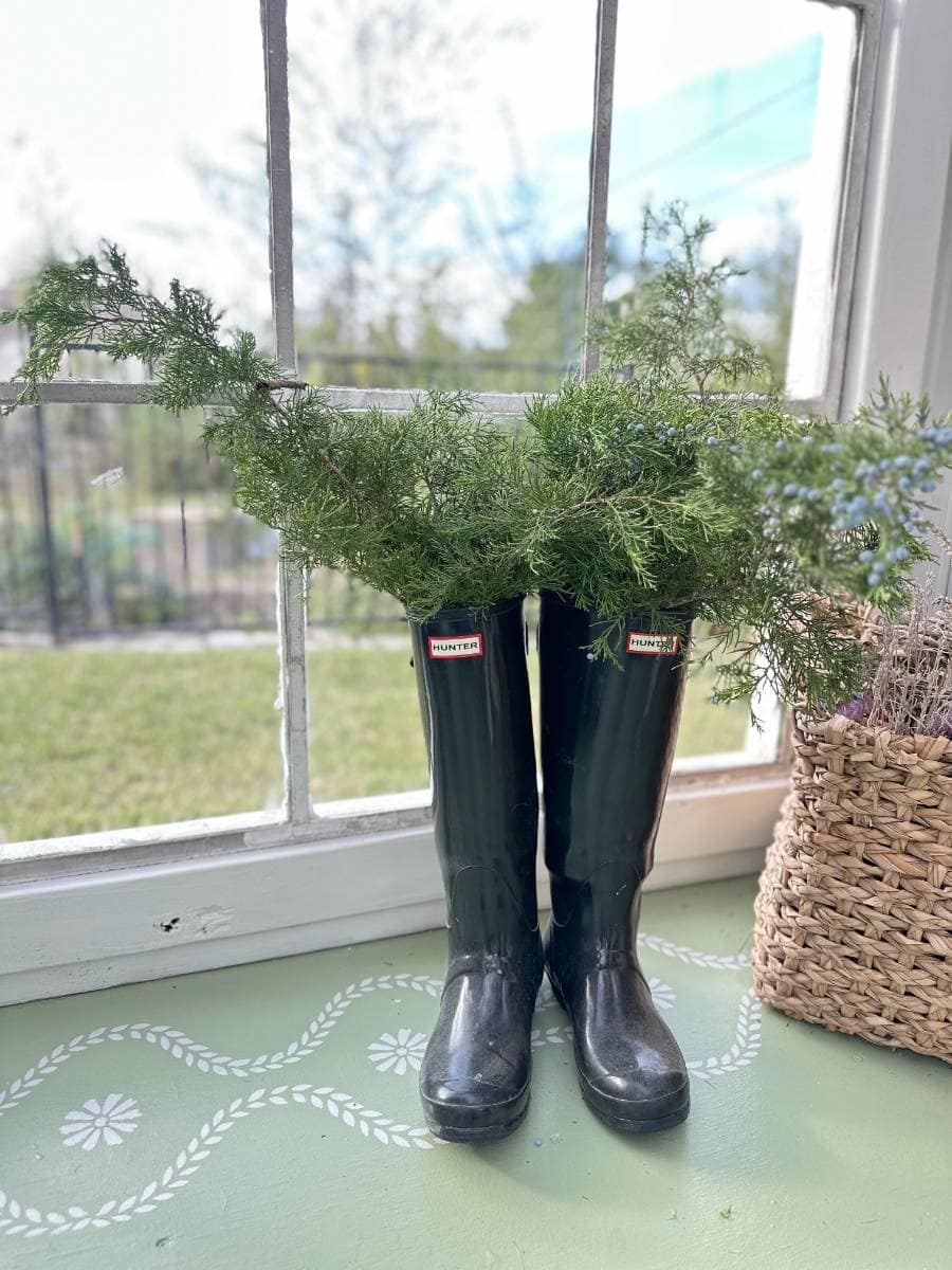 A pair of black rain boots filled with green foliage sit on a green windowsill next to a woven basket, offering a glimpse into the cozy charm of the Christmas Tour of the Garden House, with the garden visible through the window behind them.