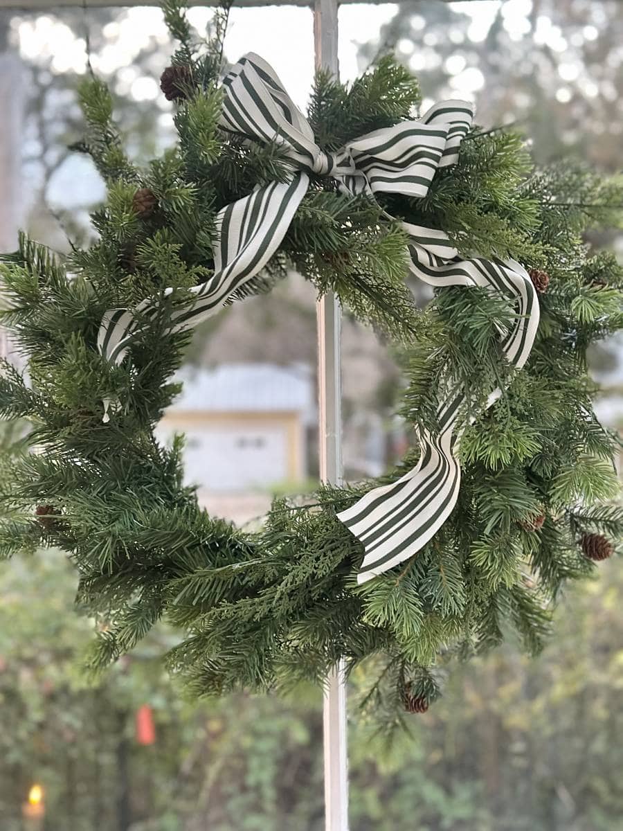 A green pine wreath decorated with a large black-and-white striped bow and small pinecones hangs on a window, adding festive charm to the Christmas Tour of the Garden House, with a blurry outdoor background visible through the glass.