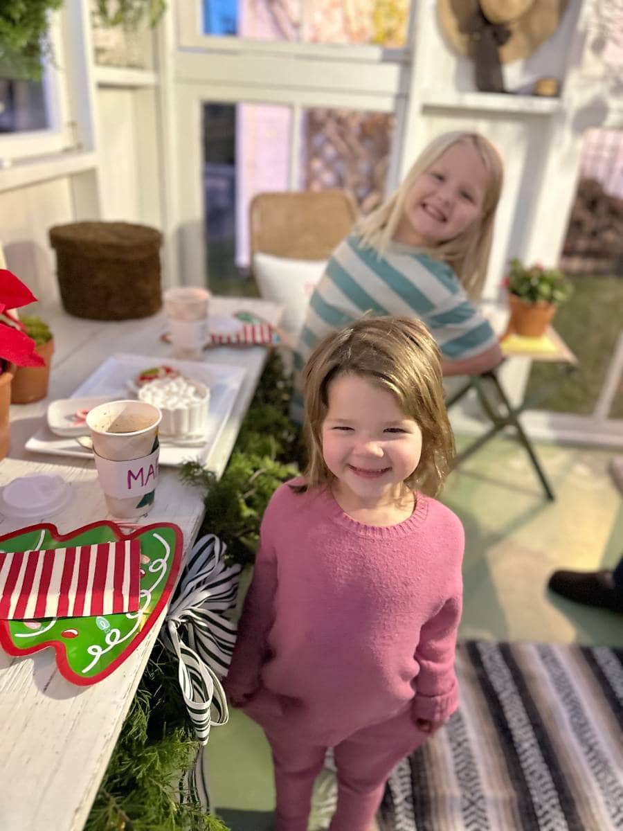 Two young children smile indoors near a table set with hot chocolate and holiday-themed plates, enjoying festive decorations after their Christmas Tour of the Garden House. One child in pink stands in the foreground, both looking happy and cheerful.