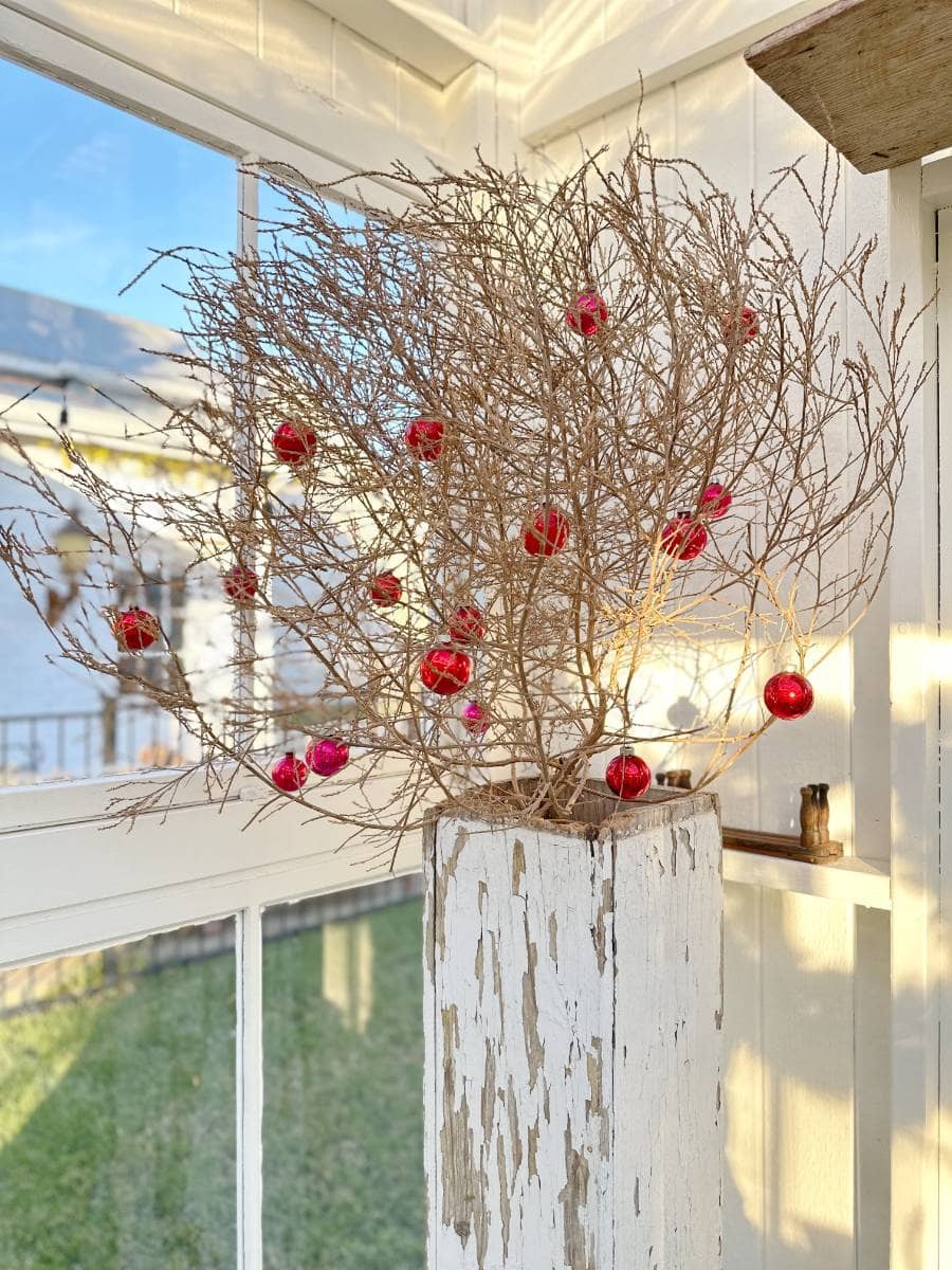 A dried, leafless branch adorned with red ornaments sits in a rustic, whitewashed wooden vase by a sunlit window—an inviting touch for the Christmas Tour of the Garden House, with a softly blurred winter scene outside.