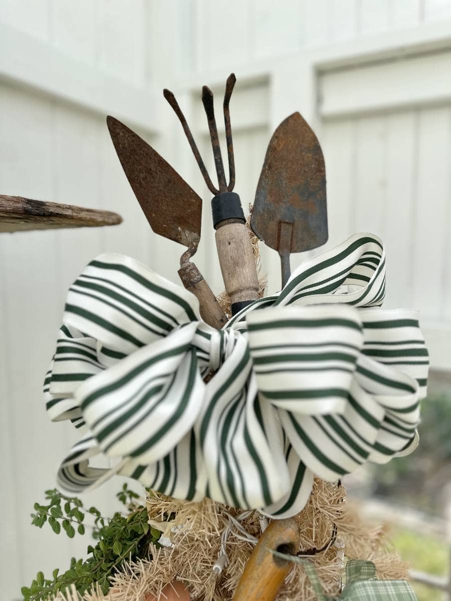 Three rusty garden tools are arranged upright behind a large white ribbon with green stripes, creating a festive touch for the Christmas Tour of the Garden House. A white wooden fence and greenery accent the background’s charm.