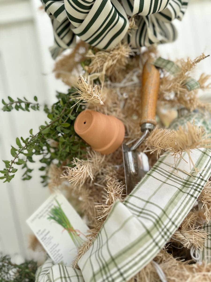 A close-up of a decorative arrangement from the Christmas Tour of the Garden House features a striped bow, a small terracotta pot, a garden trowel with a wooden handle, green foliage, and plaid ribbon against a light background.