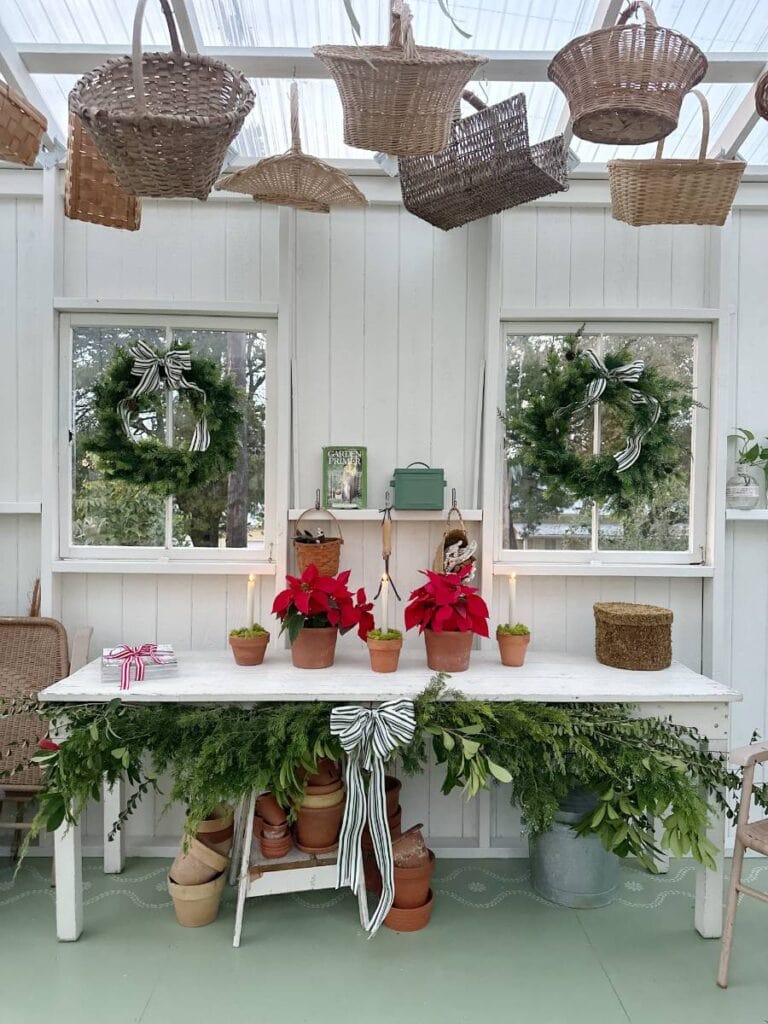 A white table with poinsettias, greenery, and clay pots stands under two windows with wreaths. Woven baskets hang from the ceiling in a bright, white room adorned for a festive Christmas Tour of the Garden House.