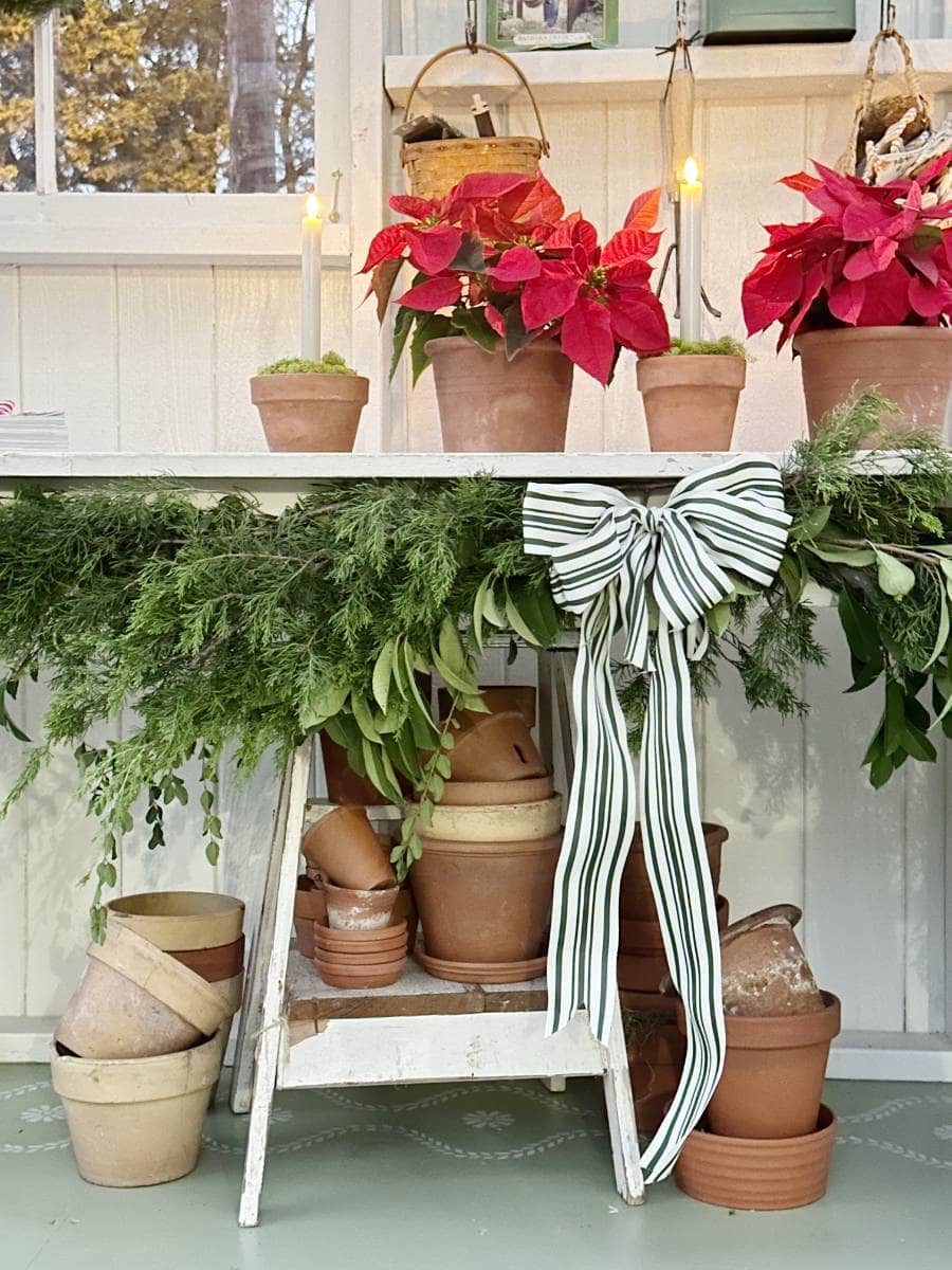 A white table decorated with a green garland and striped bow holds red poinsettias and potted plants; terracotta pots are stacked on the shelf below. Two lit candles add a warm touch to this festive Christmas Tour of the Garden House.