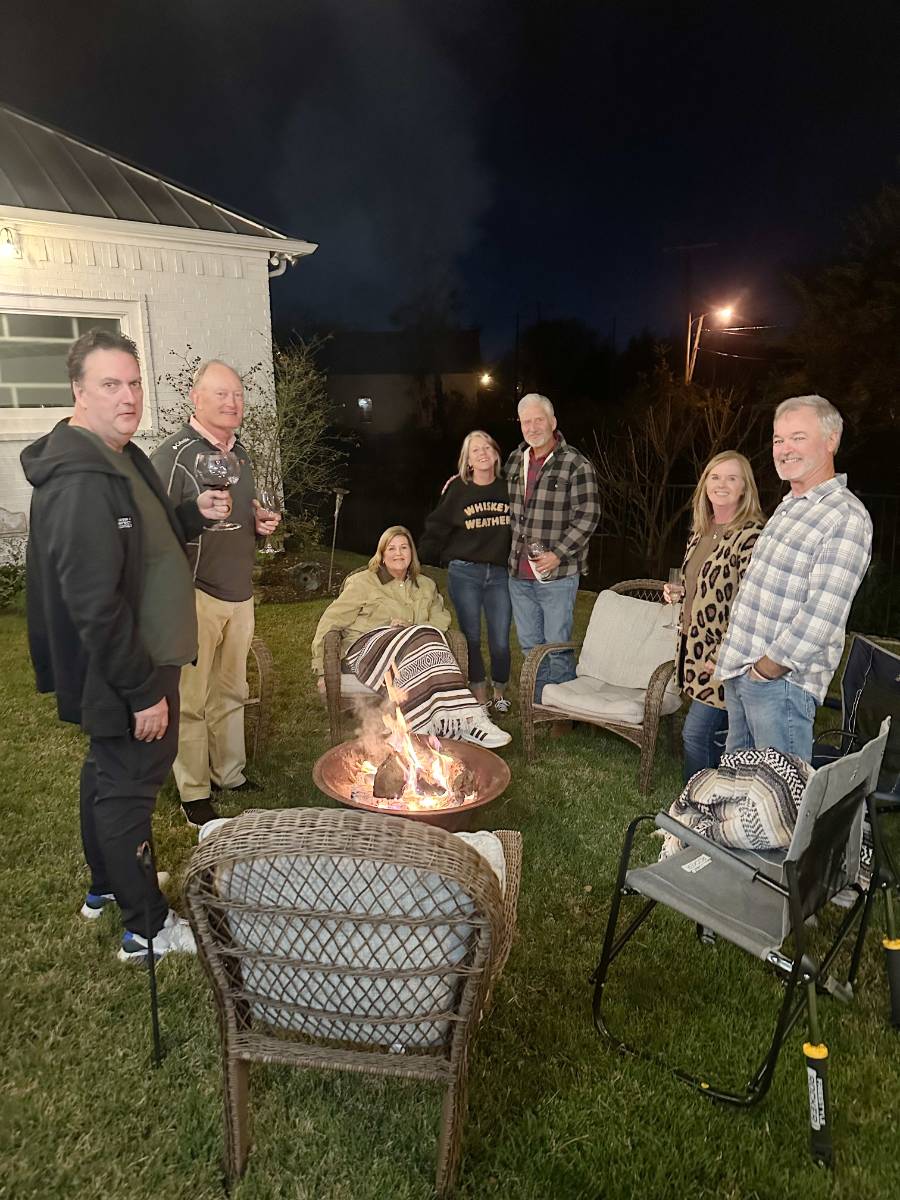 Seven adults gather around a fire pit in the backyard at night, chatting and smiling with drinks in hand. Some stand while others relax on wicker chairs, enjoying a festive atmosphere reminiscent of a Christmas Tour of the Garden House.